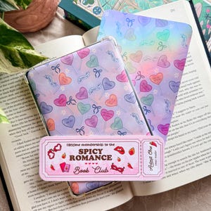 Op de afbeelding: Een verzameling leesaccessoires, waaronder een bladwijzer, een boek en een hoesje. De bladwijzer leest "lifetime membership to the SPICY ROMANCE Book Club". De hoes en bladwijzer hebben hartvormige lolly's en strikken.