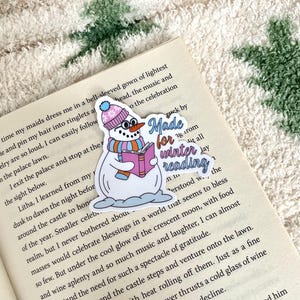 Op de afbeelding: Een grillige sticker met een sneeuwpop met een roze en blauwe hoed, een bril en een sjaal, die een roze boek vasthoudt. De tekst "Made for winter reading" staat ook op de sticker. De sticker is op een open boek geplaatst.