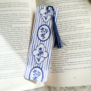 Op de afbeelding: Een witte bladwijzer met blauwe bloemen- en streepmotieven. De bladwijzer heeft een blauwe kwast en ligt op een open boek. Het ontwerp bevat ovale en wolkvormige bloemmotieven.