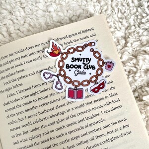 Op de afbeelding: Een sticker met de tekst "Smutty Book Club Girlie" in een kettingontwerp, met kleine illustraties van een chilipeper, handboeien, een boek, een hartvormige doos en een masker. De sticker bevindt zich op een open boek.