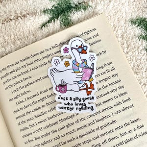 Op de afbeelding: Een witte ganzensticker met een bril, sjaal en boek, met een mok met een sneeuwvlok. De tekst luidt: "Just a silly goose who loves winter reading." De sticker staat op een boek.