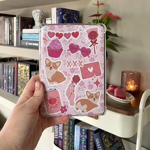 May include: A pink phone case with a variety of stickers, including a cupcake, sunglasses, hearts, a dog, a can of soda, and a rose. The case is decorated with a floral pattern.