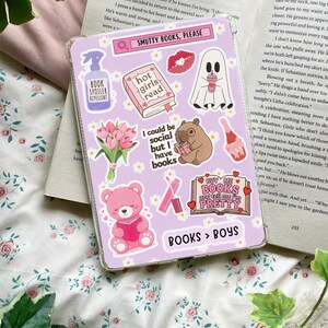 Op de afbeelding: Een paarse en witte telefoonhoes met verschillende stickers. De stickers tonen een geest, een boek met de tekst "hot girls read", een boeket roze tulpen, een beer die een boek leest, een wombat die een boek leest, een roze lippenstift en een boek met de tekst "Buy me books and tell me I'm pretty". De telefoonhoes heeft ook de tekst "Smutty Books, Please" en "Books > Boys".