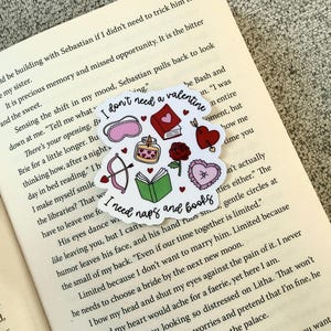 Op de afbeelding: Een sticker met de tekst "I don't need a valentine, I need naps and books" en Valentijnsdag-thema-illustraties, waaronder een hart, pijl en boog en een boek. De sticker is geplaatst op een open boek.
