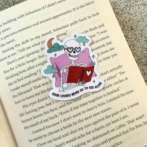 Könnte beinhalten: Ein Aufkleber mit einem Skelett mit herzförmiger Brille, das ein Buch liest. Der Aufkleber befindet sich auf einem rosa Kissen mit Mond und Wolken. Der Text lautet "BOOK LOVERS NEVER GO TO BED ALONE".