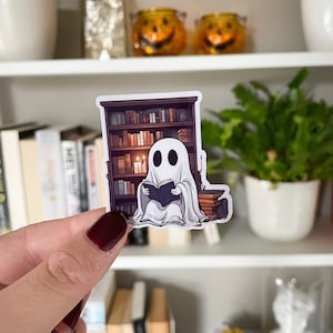 Op de afbeelding: Een sticker met een cartoongeest die een boek leest voor een boekenplank. De geest is wit met zwarte ogen en is gehuld in een wit laken. De boekenplank is bruin en gevuld met boeken.