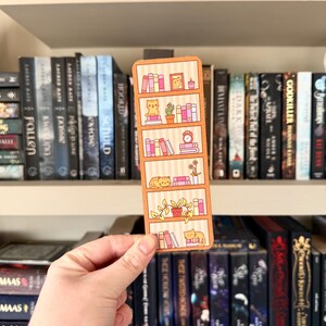 Può includere: Un segnalibro rettangolare con un design a libreria con accenti arancioni e rosa. Il segnalibro raffigura scaffali con libri, piante e gatti che dormono. Lo sfondo mostra una vera libreria con vari titoli di libri.