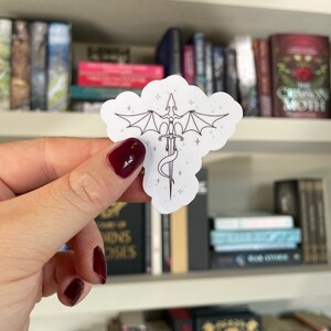 Op de afbeelding: Een witte sticker met een lijntekening van een draak die om een zwaard is gewikkeld. De draak heeft zijn vleugels uitgespreid en er zijn kleine sterren omheen.