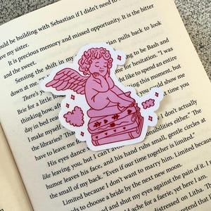 Op de afbeelding: Een roze sticker met een cherubijn met vleugels, rustend op een stapel boeken. De cherubijn bevindt zich in een ontspannen pose, met gesloten ogen. De sticker heeft een witte rand en is geplaatst op een open boek.