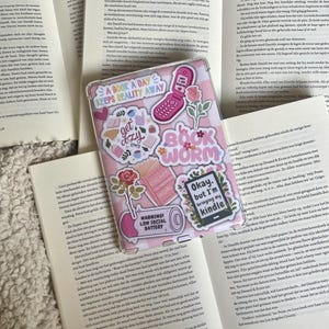 Op de afbeelding: Een roze en wit boekjournaal met verschillende stickers. De stickers bevatten een telefoon, een boekenworm, een waarschuwingsbord en een 'get cozy'-sticker. Het journaal heeft de tekst 'A Book A Day Keeps Reality Away' op de voorkant.