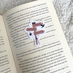 Op de afbeelding: Een close-up van een boekpagina met tekst over een boottocht. Een sticker met een bruine achtergrond en witte tekst toont de namen van drie fictieve steden: Velaris, Terrasen en Crescent City. De sticker heeft een kroon en een draakontwerp.