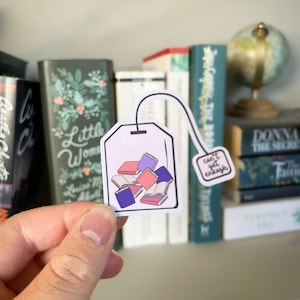 Op de afbeelding: Een witte theezakje sticker met een stapel kleurrijke boeken erin en een klein label met de tekst "can't get enough".