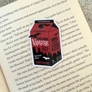 Op de afbeelding: Een sticker met een rode verpakking van "Vampire Aid" met de tekst "Fresh Blood" en "100% Vegan Human Blood". Het ontwerp bevat vleermuizen en de zin "Bloody Delicious".
