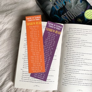 Può includere: Un libro aperto con due segnalibri, uno arancione e uno viola, con l'etichetta "Empire of Storms Tower of Dawn Tandem Read". Il libro è di Sarah Maas. La copertina del libro è visibile.