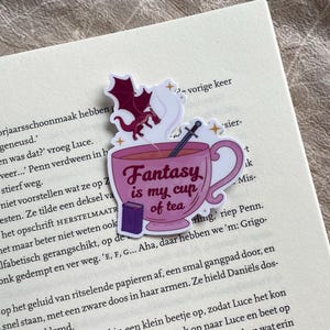 Op de afbeelding: Een roze theekop-sticker met een draak en een zwaard erin, met de tekst "Fantasy is my cup of tea". De sticker is geplaatst op een boekpagina met tekst over een personage genaamd Penn.