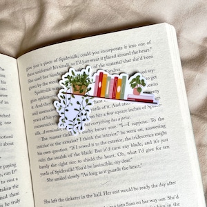 Op de afbeelding: Een boekenlegger met een bloemenontwerp en een stapel kleurrijke boeken. De boekenlegger toont een plant in een pot met groene bladeren en een kleine pot. De boeken zijn verticaal gerangschikt en hebben verschillende gekleurde ruggen. De boekenlegger is geplaatst op een pagina van een boek met tekst over spinzijde.