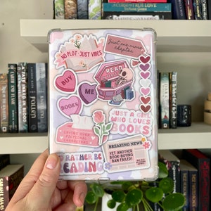 Könnte beinhalten: Eine transparente Tablet-Hülle, verziert mit Aufklebern. Die Aufkleber zeigen Sätze wie „No Plot Just Vibes“, „Read in Peace“ und „Just a Girl Who Loves Books“. Weitere Aufkleber zeigen Herzen, Bücher und eine Schlagzeile.