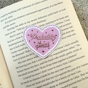 May include: A pink heart-shaped sticker with the words "Probably Smut" in a cursive font. The sticker has a white scalloped border and small heart accents. The sticker is placed on top of an open book.