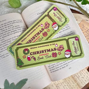 Può includere: Due segnalibri verdi e dorati per l'iscrizione al Christmas Book Club con illustrazioni festive di Babbo Natale, un albero di Natale e un cavallo a dondolo. I segnalibri sono appoggiati su un libro aperto.