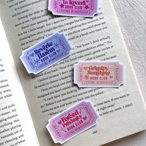Puede incluir: Cuatro pegatinas coloridas con forma de boleto con texto, incluyendo "Friends to Lovers Book Club" y "Reverse Harem Book Club", se muestran en un libro abierto. Las pegatinas tienen una estética vintage con diseños florales y de corazón.