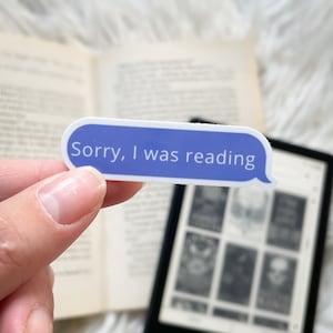 Op de afbeelding: Een blauwe en witte sticker met de tekst "Sorry, I was reading" op een witte achtergrond. De sticker wordt vastgehouden door een hand voor een boek en een e-reader.