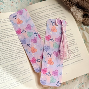 Può includere: Due segnalibri a forma di lecca lecca a forma di cuore con sfondo lilla chiaro. I segnalibri presentano lecca lecca a cuore colorati, fiocchi e la scritta "Sucker For". Una nappa rosa pende da un segnalibro.