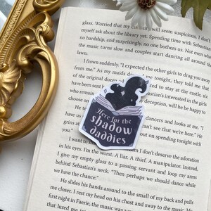 Op de afbeelding: Een zwart-witte sticker met een boek en een schaduwfiguur. De sticker zegt "Here for the shadow daddies".