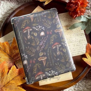 Op de afbeelding: Een doorzichtige beschermhoes voor een e-reader met een donkere achtergrond met een patroon van paddenstoelen, varens en bessen. De hoes ligt op een houten dienblad met herfstbladeren en oranje bloemen.