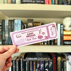 Könnte beinhalten: Ein rosa Lesezeichen mit dem Text "lifetime membership to the ANTI SOCIAL Book Club" und Illustrationen eines Buches, einer Teekanne und einer Kaffeetasse. Das Lesezeichen wird vor einem Bücherregal mit Büchern gehalten.