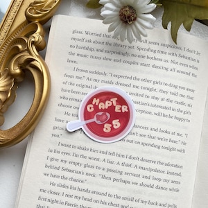 Può includere: Un adesivo rosso e bianco con il testo "Capitolo 55" in un cerchio bianco su sfondo rosso. L'adesivo è posizionato su una pagina di un libro con testo su un personaggio di nome Sebastian.