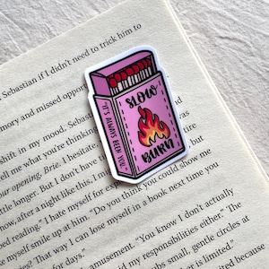 Op de afbeelding: Roze sticker van een luciferdoosje met de woorden "SLOW BURN" en een vlammenafbeelding. De sticker heeft ook de tekst "IT'S ALWAYS BEEN YOU." Het is geplaatst op een open boek.