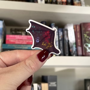 Può includere: Un adesivo con un design ad ala di pipistrello viola scuro con schizzi colorati e un'impronta di mano. L'adesivo ha un bordo bianco e viene tenuto davanti a una libreria piena di libri.