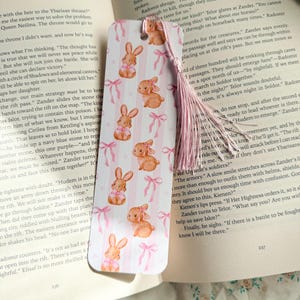 Può includere: Un segnalibro stravagante con un motivo ripetuto di coniglietti ad acquerello con fiocchi rosa su uno sfondo a righe bianche e rosa. Una nappa rosa è appesa.