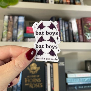 Op de afbeelding: Een witte sticker met de tekst "bat boys" en vleermuisvleugel illustraties in donkerpaars. De sticker bevat ook de zin "watcha gonna do" en kleine steraccenten. De sticker wordt voor een boekenplank gehouden.