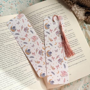 Può includere: Un set di segnalibri a tema fantasy e una custodia per libri abbinata. I segnalibri e la custodia presentano un motivo ripetuto di draghi, lune, chiavi, fiocchi e piume in morbidi colori pastello. Un nappa rosa pende da un segnalibro.