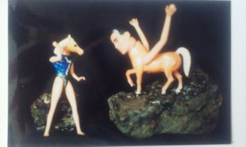 May include: A toy horse with a human head and a toy person with a horse head stand on a rocky surface. The horse is brown and white, and the person is wearing a blue swimsuit.