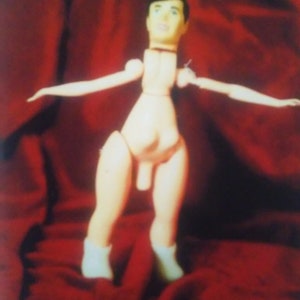 Puede incluir: Una muñeca articulada de madera con un tono de piel claro, que lleva botas blancas. La muñeca tiene un físico masculino y posa con los brazos extendidos. El fondo es una tela de color rojo oscuro.