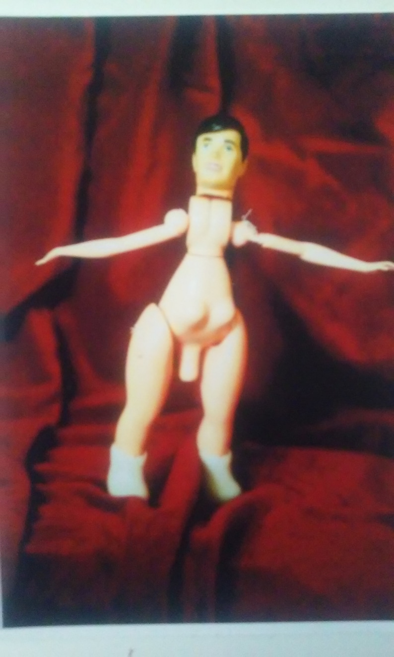 May include: A wooden articulated doll with a light skin tone, wearing white boots. The doll has a male physique and is posed with arms outstretched. The background is a deep red fabric.