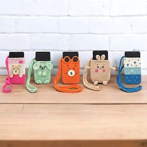 Könnte beinhalten: Fünf farbenfrohe, gehäkelte Handyhüllen mit Tier- und geometrischen Designs. Die Hüllen sind in Pink, Grün, Orange, Beige und Blau gehalten, jede mit einem passenden Riemen. Die Hüllen zeigen ein Bären-, Hunde-, Bären-, Hasen- und Sternmuster.
