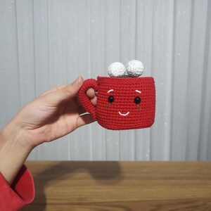 Op de afbeelding: Een handgemaakte rode gehaakte mok met een lachend gezicht en twee witte pompons bovenop. De mok heeft een handvat en zwarte ogen. De mok is een schattig, grillig ontwerp.
