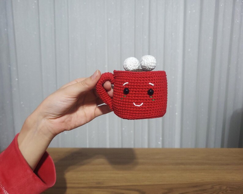 Op de afbeelding: Een handgemaakte rode gehaakte mok met een lachend gezicht en twee witte pompons bovenop. De mok heeft een handvat en zwarte ogen. De mok is een schattig, grillig ontwerp.