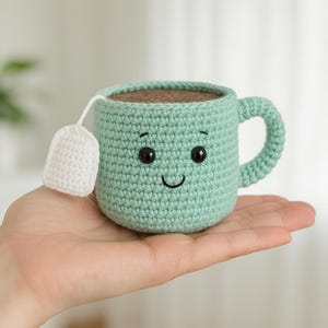 Peut inclure: Une tasse à thé en crochet de couleur verte avec un visage souriant et des yeux noirs. La tasse à thé a un sachet de thé en crochet blanc attaché sur le côté.