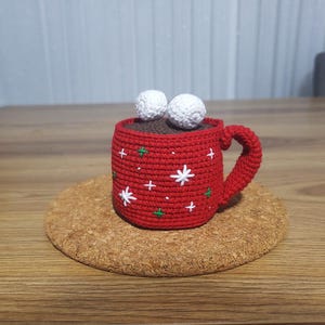 Op de afbeelding: Een gehaakte rode mok met een bruin interieur, witte ster- en groene kruisaccenten en twee witte marshmallow-achtige ballen bovenop. De mok staat op een ronde kurken onderzetter.