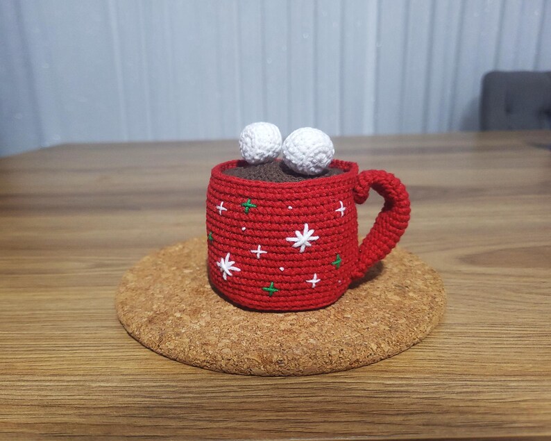 Op de afbeelding: Een gehaakte rode mok met een bruin interieur, witte ster- en groene kruisaccenten en twee witte marshmallow-achtige ballen bovenop. De mok staat op een ronde kurken onderzetter.