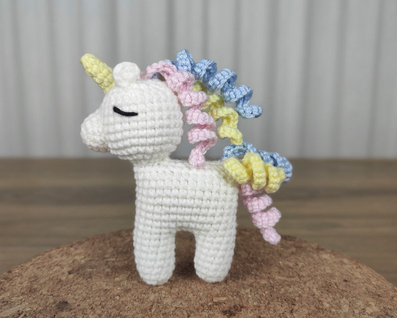 May include: A white crocheted unicorn with a pink, yellow, and blue curly mane and tail.