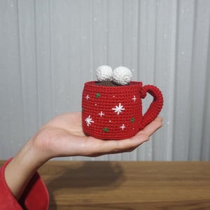 Op de afbeelding: Een handgemaakte rode gehaakte mok met witte sneeuwvlokken en groene kruissteekdetails. De mok is gevuld met bruin materiaal en afgewerkt met twee witte pompons. De mok wordt in een hand gehouden.
