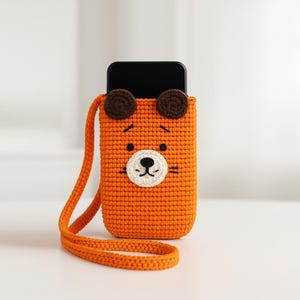 May include: Orange crocheted phone case with a bear face design. The case has a brown strap and black eyes.