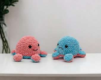 May include: Two handmade crochet octopus plushies. One is pink and blue with a smiling face, the other is blue and pink with a sad face. The plushies are sitting on a white surface.