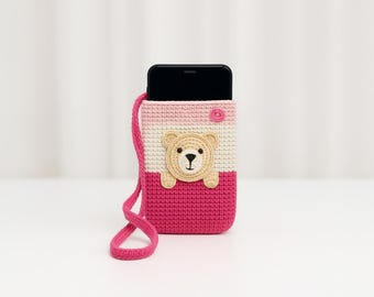 Puede incluir: Una funda de teléfono de crochet rosa y blanca con una cara de oso en la parte delantera. La funda tiene una correa rosa.