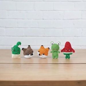Puede incluir: Una colección de cinco figuras amigurumi de ganchillo hechas a mano. Las figuras incluyen un cactus verde con flores rosas, un zorro marrón y blanco, un zorro naranja y blanco, una rana verde y una sandía roja con detalles verdes.